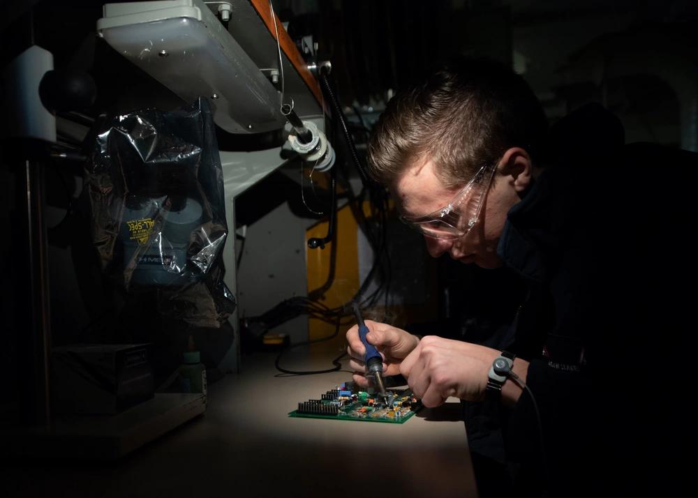 Real photo: aviation electronics technician soldering a circuit board at a bench under a work lamp